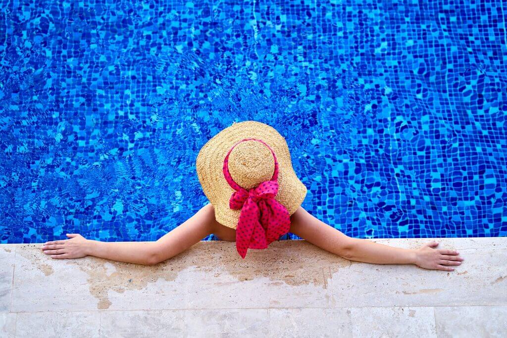 A woman in a hat and scarf sitting by a swimming pool
